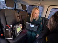 Ambulance crew speaking to a female patient in the rear of a  Mental Health Response Vehicle. The staff member is holding a laptop, next to her is a piece of equipment labelled 'Corpuls'